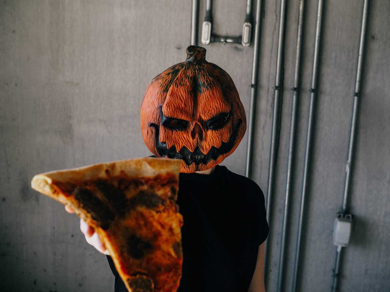 Man wearing a spooky Jack-o-lantern mask holding a pizza 