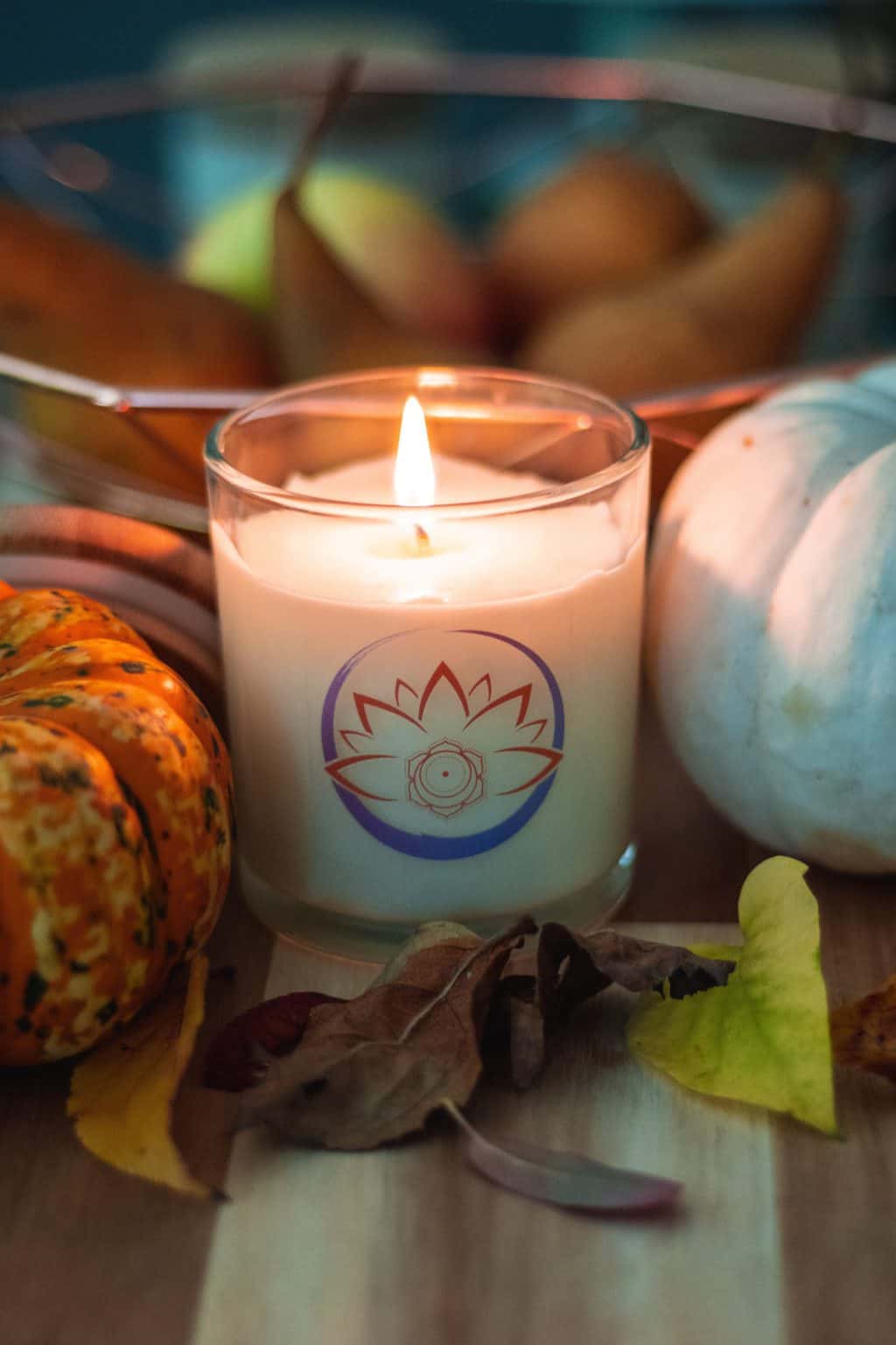A white lighted scented candle displayed among pumpkins and pears