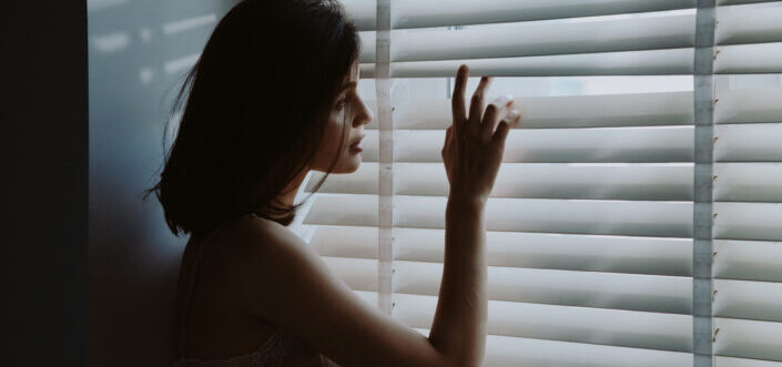 Woman in white nightdress looking through window blinds