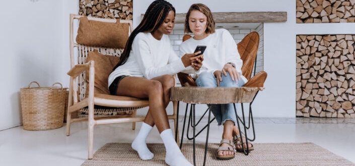 Two ladies looking at a phone while sitting on wooden chairs